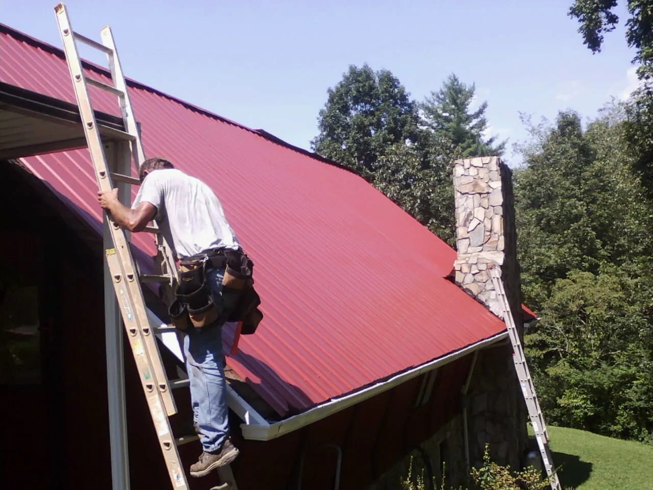 Roofer on a ladder beside a red metal roof and stone chimney — Soffit & Fascia Repair by Roofing Brothers in Mount Enterprise